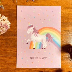Puede incluir: Una tarjeta rosa con un unicornio blanco de pie sobre un arcoíris. El unicornio tiene una melena y una cola moradas. La tarjeta tiene un fondo rosa con confeti de colores y el texto "QUEER MAGIC".