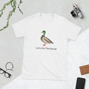 Può includere: Maglietta bianca con un'immagine di un'anatra con la testa verde e il testo "Let's Get This Bread".