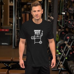 Könnte beinhalten: Ein schwarzes T-Shirt mit einer weißen Grafik eines Grills mit Rauch aus dem Schornstein. Der Text "Smokin' Meats" befindet sich über dem Grill.
