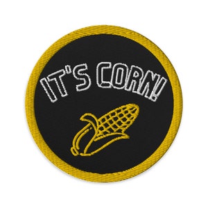 Può includere: Una toppa rotonda nera ricamata con un bordo giallo. La toppa presenta il testo "IT'S CORN!" in bianco e un contorno giallo di una pannocchia di mais.