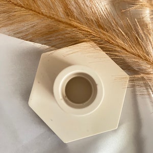 May include: White hexagonal ceramic candle holder with a round opening in the center. The holder is on a white fabric surface.
