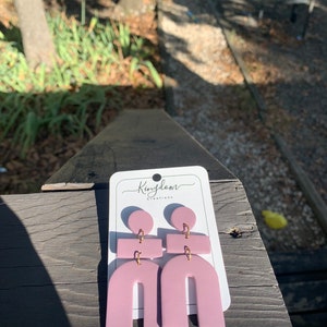 May include: A pair of pink geometric earrings with gold accents. The earrings are shaped like arches and have a small rectangle and circle on top. The earrings are on a white card with the text "Kingdom Creations".
