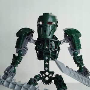 Op de afbeelding: Een groen en grijs gearticuleerd robot speelgoed met een helm en twee zwaarden. De robot staat in een vechtpose.