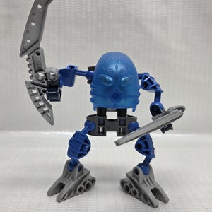 Puede incluir: Una figura de acción azul y gris con una cabeza redondeada y dos armas plateadas. La figura tiene extremidades y pies azules y está posada como en acción. La figura está sobre una superficie blanca.