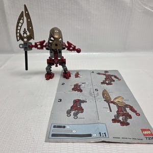 Op de afbeelding: Een Lego Bionicle-figuur met een gouden masker en rode en grijze lichaamsdelen, met een gouden en zwarte speer. Een Lego instructievel ligt op de voorgrond.