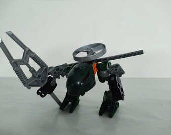 以下が含まれることがあります： 回転するプロペラと大きな可動式アームを持つ緑と灰色のオモチャのロボット。ロボットの頭には黒とオレンジのデザインが施されています。
