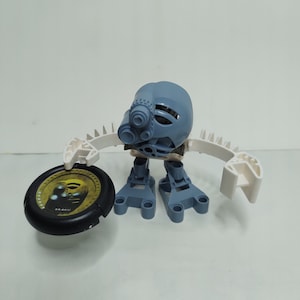 May include: A blue and white Lego robot with a black disc. The disc has a yellow and black design with the word "Araku" on it.