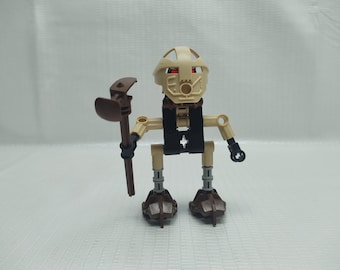 以下が含まれることがあります： 赤い目をしたベージュと茶色のレゴミニフィギュアで、茶色の杖を持っています。ミニフィギュアは黒いベストを着ており、胸に黒い十字架のあるベージュの頭をしています。