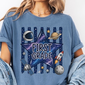 May include: A blue t-shirt featuring a large dark blue star with a galaxy print and the text FIRST GRADE. The design includes a white astronaut helmet, a planet with rings, a rocket ship, a satellite, and a moon against vertical stripes.