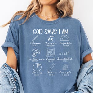 May include: A blue t-shirt with the text "GOD SAYS I AM" and various words and symbols, including "Chosen," "Unique," and "Capable." The shirt is made of a soft material and has rolled-up sleeves. A denim jacket is draped over the shoulders.