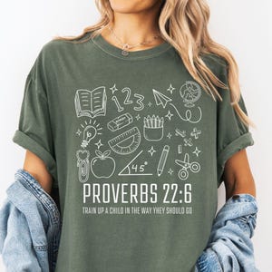 May include: Moss green t-shirt with a white graphic featuring school-related illustrations and the text "PROVERBS 22:6 TRAIN UP A CHILD IN THE WAY THEY SHOULD GO." The design includes an open book, numbers, a paper airplane, a globe, and other school supplies.