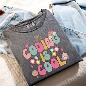 May include: A dark gray t-shirt with the text "Coding is Cool" in colorful, retro-style lettering. The design includes a rainbow, stars, gears, and a backpack.