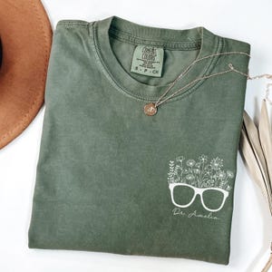 May include: A green t-shirt with a white graphic of a pair of glasses with flowers growing out of them. The text "Dr. Amelia" is below the glasses.