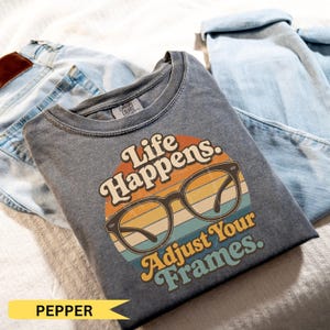 May include: A charcoal gray crew neck t-shirt featuring a graphic of black eyeglasses centered over a circular sunset design with horizontal stripes in orange, yellow, and teal. The text reads Life Happens. Adjust Your Frames. The shirt has a tag.