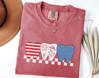 May include: Red, white, and blue tooth graphic on a red t-shirt. The teeth are decorated with stars and fireworks. The shirt has a distressed look and is made of a soft, comfortable fabric.