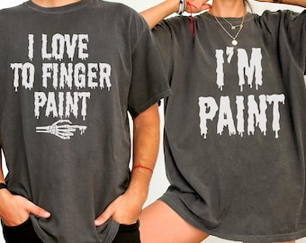 Op de afbeelding: Twee donkergrijze t-shirts met witte tekst. Eén shirt zegt "I LOVE TO FINGER PAINT" met een skelet hand grafiek. De andere zegt "I'M PAINT". Beide shirts hebben een verweerde, vintage uitstraling.