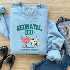 May include: Light blue sweatshirt with a green graphic of a heart and a coffee cup with the text "Neonatal ICU" and "Guardians of Hope" below. The text says "Where love, like a gentle breeze, cradles fragile beginnings, helping tiny miracles to unfold."