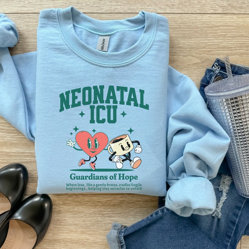 May include: Light blue sweatshirt with a green graphic of a heart and a coffee cup with the text "Neonatal ICU" and "Guardians of Hope" below. The text says "Where love, like a gentle breeze, cradles fragile beginnings, helping tiny miracles to unfold."