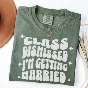May include: A green t-shirt with white text that reads 'Class Dismissed I'm Getting Married' in a retro font. The shirt has a distressed look and is folded in half. A gold necklace is draped over the shirt.