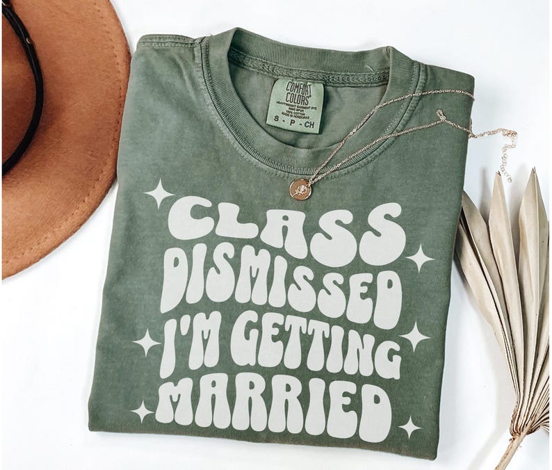 May include: A green t-shirt with white text that reads 'Class Dismissed I'm Getting Married' in a retro font. The shirt has a distressed look and is folded in half. A gold necklace is draped over the shirt.