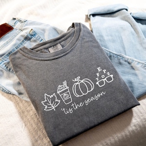 May include: A charcoal gray t-shirt with a white graphic of a fall leaf, pumpkin spice latte, pumpkin, glasses, and the text "'tis the season". The shirt is folded next to a pair of light blue jeans.