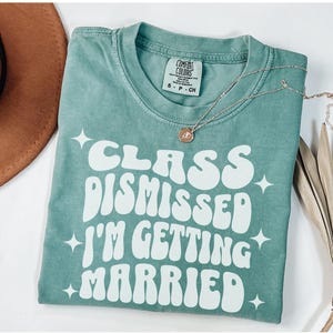 May include: A teal blue t-shirt with white text that reads 'Class Dismissed I'm Getting Married' in a retro font. The text is surrounded by small white stars.