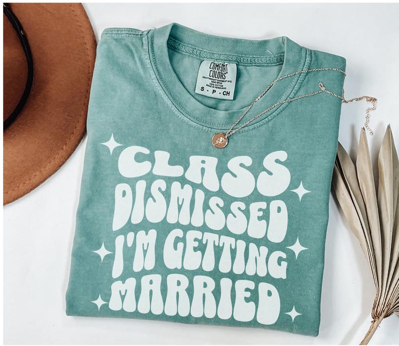 May include: A teal blue t-shirt with white text that reads 'Class Dismissed I'm Getting Married' in a retro font. The text is surrounded by small white stars.