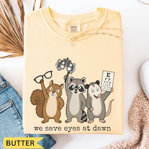 May include: A butter yellow t-shirt featuring a graphic of a squirrel holding glasses, a raccoon holding an ophthalmic phoropter, and an opossum holding an eye chart. The text below the animals reads, "we save eyes at dawn" in black font.