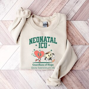 May include: A beige sweatshirt with a green graphic design that says "Neonatal ICU" with a heart and coffee cup illustration. The text below the graphic says "Guardians of Hope" and "Where love, like a gentle breeze, cradles fragile beginnings, helping tiny miracles to unfold."