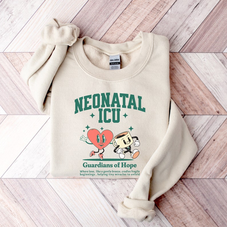 May include: A beige sweatshirt with a green graphic design that says "Neonatal ICU" with a heart and coffee cup illustration. The text below the graphic says "Guardians of Hope" and "Where love, like a gentle breeze, cradles fragile beginnings, helping tiny miracles to unfold."