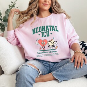 May include: Pink sweatshirt with a green graphic design that says "Neonatal ICU" and features a heart and a coffee cup with the text "Guardians of Hope" below. The text says "Where love, like a gentle breeze, cradles fragile beginnings, helping tiny miracles to unfold."
