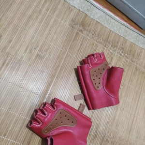May include: A pair of red fingerless gloves with brown leather accents. The gloves have a perforated brown leather patch on the back of the hand and white stitching. A small brown strap is on the wrist.