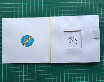 Puede incluir: Tarjeta blanca abierta con una ilustración de balón azul y amarillo en el lado izquierdo. A la derecha, una ilustración de una mano con el dedo índice apuntando hacia arriba. La tarjeta está pegada con cinta adhesiva blanca.