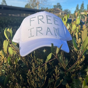 Op de afbeelding: Witte truckerpet met de tekst "FREE IRAN" in zilveren strass-steentjes. De pet heeft een mesh achterkant en een gebogen klep. De pet wordt buiten getoond, omringd door groen gebladerte.