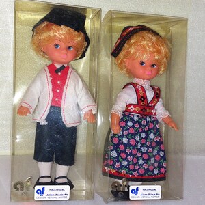 Peut inclure: Deux poupées en costumes traditionnels norvégiens, l'une portant un chapeau noir et une chemise blanche avec des bordures rouges, l'autre portant une robe rouge et blanche à imprimé floral. Les deux poupées sont emballées dans des boîtes en plastique transparent avec le texte "Hallingsdal Allan Flink As Design Verdal Norge".