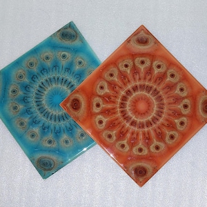 Op de afbeelding: Twee vierkante onderzetters met een glanzende afwerking. Eén is turquoise blauw, de andere is oranje. Beide hebben een centraal cirkelvormig ontwerp met een herhalend patroon van oogachtige vormen in bruin- en beigetinten. De onderzetters zijn waarschijnlijk gemaakt van hars.