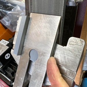 May include: A close-up of a metal sharpening jig with a grey rubber block attached to the side. The jig is designed to hold a knife blade at a specific angle for sharpening.