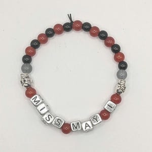 May include: A beaded bracelet with red, black, and gray beads. The bracelet features silver skull charms and square letter beads spelling "MISS MAY". The beads are strung on a black elastic cord.