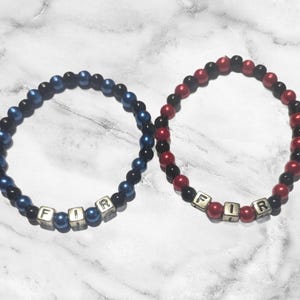 May include: Two beaded bracelets with black and blue beads and black and red beads. The bracelets have silver letter beads spelling out the word "FIR".