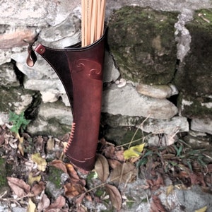May include: A brown leather quiver with a decorative design holds a bundle of wooden arrows with red and black fletching.