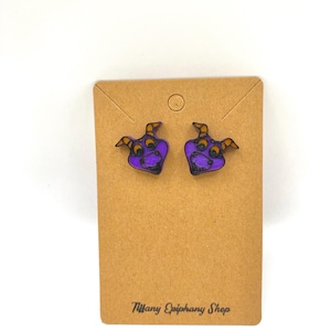 May include: A pair of purple and black earrings shaped like a cartoon character's face with orange accents. The earrings are displayed on a brown card with the text "Tiffany Epiphany Shop".
