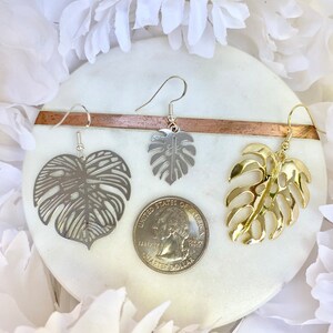 May include: Three pairs of earrings featuring a monstera leaf design. One pair is silver, one is gold, and one is a silver tone with a cut-out design. The earrings are displayed on a white marble surface with a US quarter for scale.