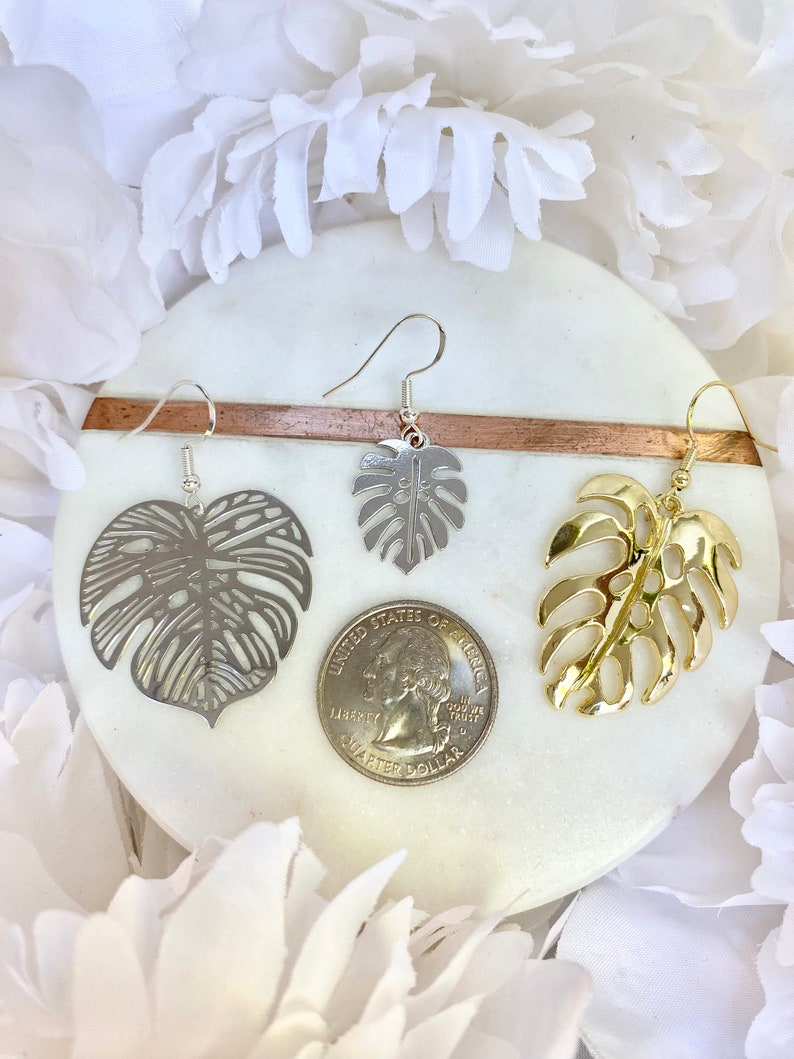 May include: Three pairs of earrings featuring a monstera leaf design. One pair is silver, one is gold, and one is a silver tone with a cut-out design. The earrings are displayed on a white marble surface with a US quarter for scale.