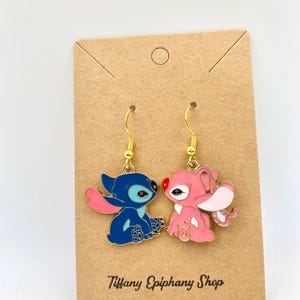 Op de afbeelding: Een paar goudkleurige oorhangers met blauwe en roze stripfiguren. Het blauwe figuur heeft grote oren en een roze arm, terwijl het roze figuur grote oren en een rode neus heeft. De oorbellen zitten op een bruine kaart met de tekst "Tiffany Epiphany Shop".