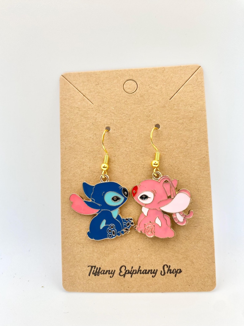 Op de afbeelding: Een paar goudkleurige oorhangers met blauwe en roze stripfiguren. Het blauwe figuur heeft grote oren en een roze arm, terwijl het roze figuur grote oren en een rode neus heeft. De oorbellen zitten op een bruine kaart met de tekst "Tiffany Epiphany Shop".