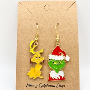 Peut inclure: Boucles d'oreilles pendantes dorées représentant des personnages de dessins animés. Une boucle d'oreille représente un chien jaune avec des bois, l'autre un personnage vert avec un bonnet de Père Noël. Les boucles sont sur une carte marron avec le texte "Tiffany Epiphany Shop."