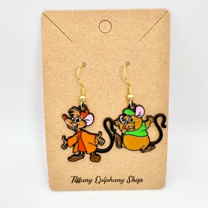 Könnte beinhalten: Ohrringe mit Cartoon-Maus-Charakteren in Orange, Grün und Braun. Die Ohrringe sind an goldfarbenen Fischhaken-Ohrdrähten befestigt und werden auf einer braunen Karte mit dem Text "Tiffany Epiphany Shop" präsentiert.