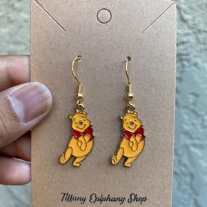 May include: A pair of gold-toned dangle earrings featuring yellow and red enamel Winnie the Pooh charms. The earrings are attached to a rectangular brown cardboard display card with the text Tiffany Epiphany Shop printed at the bottom.