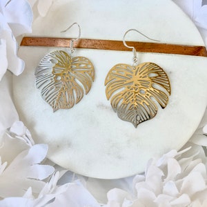 May include: A pair of gold and silver earrings featuring a delicate, cutout monstera leaf design. The earrings are displayed on a white marble surface.