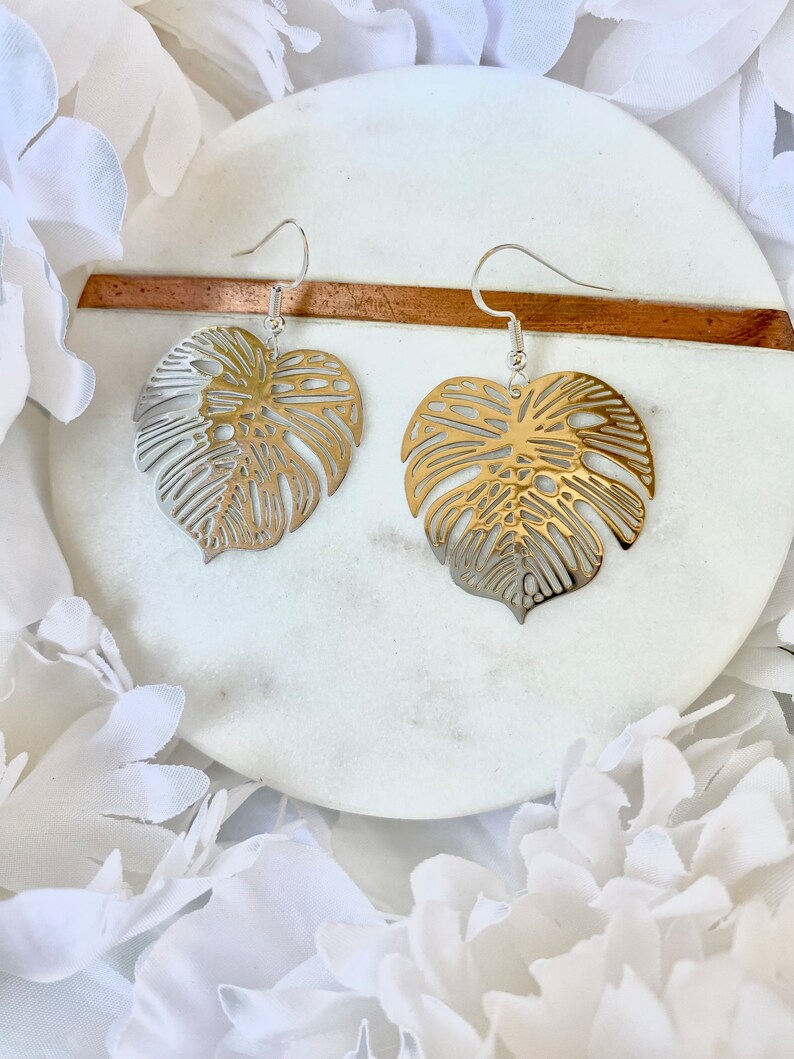 May include: A pair of gold and silver earrings featuring a delicate, cutout monstera leaf design. The earrings are displayed on a white marble surface.